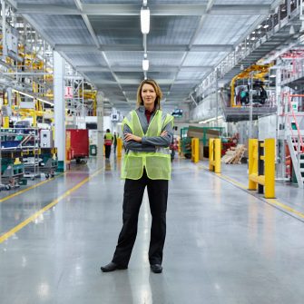 Portrait of mature female supervisor standing with arms crossed at automobile industry. Full length of confident manager is working at factory. She is wearing reflective clothing.