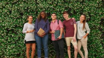 five students stand against a green backdrop smiling