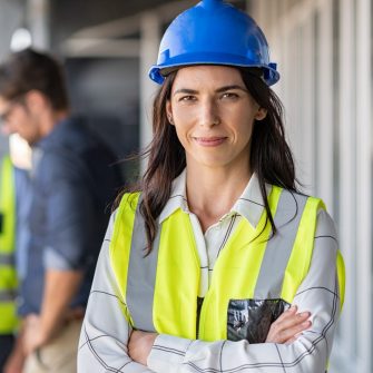 Portrait of woman engineer at building site looking at camera with copy space. Mature construction manager standing in yellow safety vest and blue hardhat with crossed arms. Successful confident architect at construction site with team discussing in background.