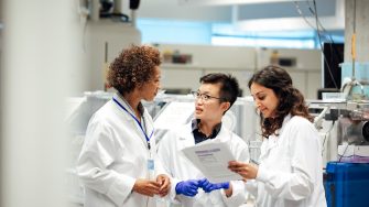 Diverse mix of technicians, wearing lab coats, debate working procedure, in a disease research facility. Nuclear magnates resonance equipment, is in the background, this high end apparatus can be used to discover information on conditions such as dementia, diabetes, autism and cancer. Focus technique and layers, ensure the group of female science graduates and manager are the centre of attention. This is a realistic scenario.