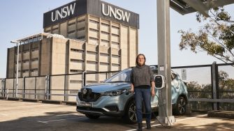 A woman stands beside a light-blue electric car plugged into a charging station on the rooftop of UNSW’s Botany Street car park.