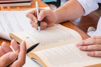 Two people studying together, one is making notes on an open book, while the other has a stack of papers they are reading from. 
