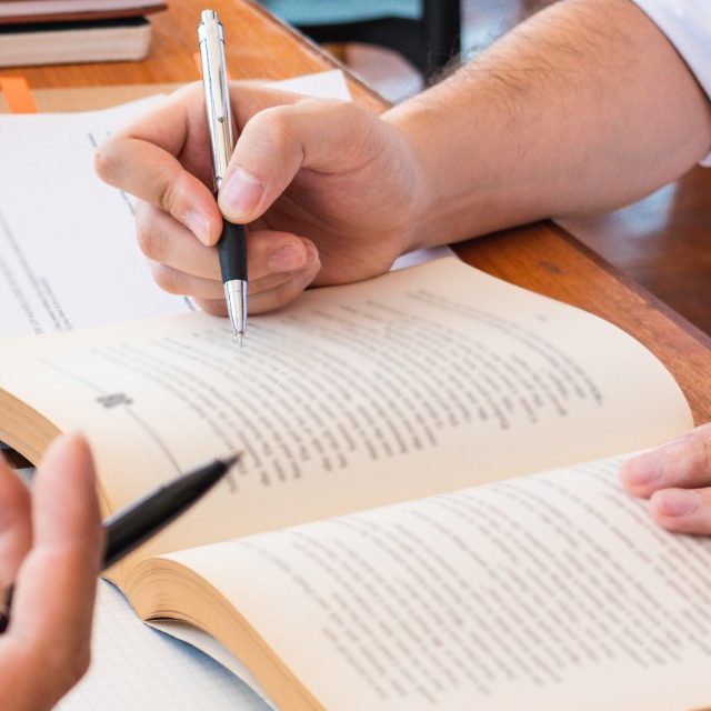 Two people studying together, one is making notes on an open book, while the other has a stack of papers they are reading from. 