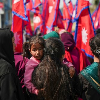 A loose group of Nepalese people congregate, one holding a child, featuring several Nepalese flags in the background.