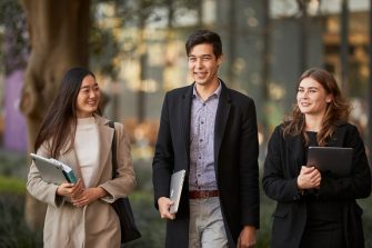 Three Law students walking in front of Law Building, dressed in smart attire, walking towards the camera, greenary and reflections in glass of law building in the background