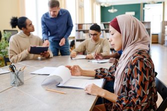 Muslim woman sitting at table looking through sheets of paper with other people studying in the background