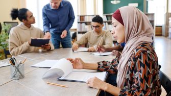 Muslim woman sitting at table looking through sheets of paper with other people studying in the background