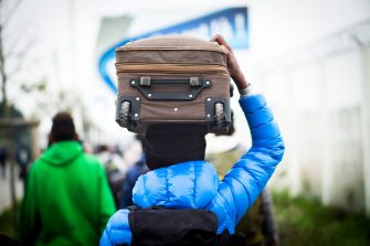 Refugee with back to camera holding a suitcase on head with one arm