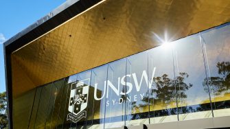 Glass front of Sir John Clancy Auditorium showing UNSW Sydney and Crest in white lettering. Gold coloured ceiling glistening and reflected in the glass.