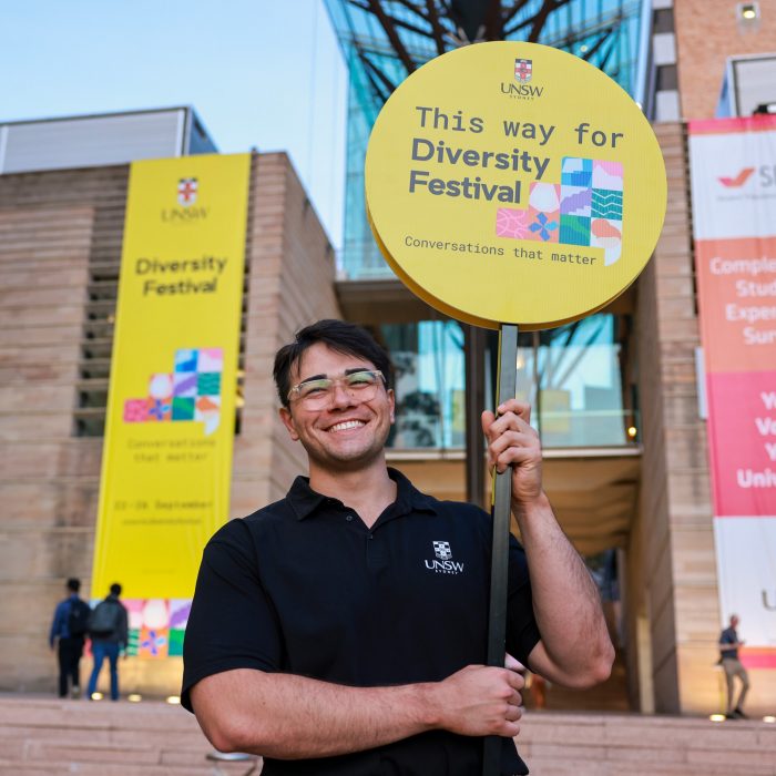 A person smiling broadly and holding a sign 'This way for Diversity Festival'. The Scientia building with a large Diversity Festival banner is in the background.