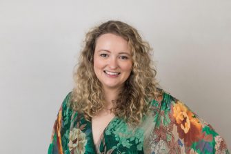 A professional headshot of a smiling individual with curly hair, wearing a colorful floral outfit. The neutral background highlights the subject's attire and friendly demeanor.