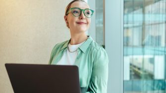 A young professional woman with glasses stands confidently holding a laptop. She exudes a sense of motivation and readiness, leaning against a glass wall in a modern office setting.