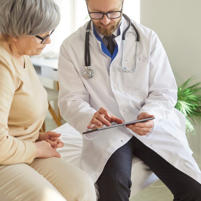 Doctor or nurse conducts medical consultation for elderly patient in hospital office, using tablet pc for care in a professional setting with focus on communication and healthcare support.