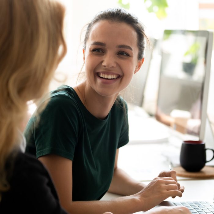 Happy millennial intern, student girl talking to teacher, consulting tutor, mentor at computer in training center, discussing new professional skills, smiling, laughing. Business education, internship
