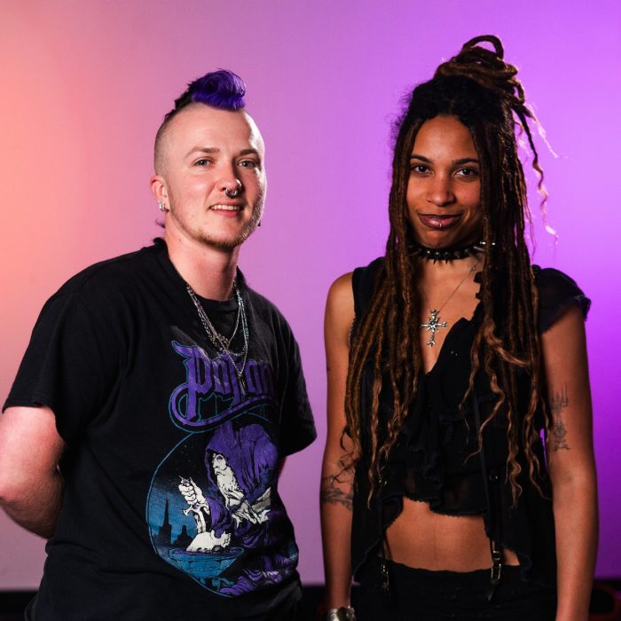 Two people are smiling to the camera against a pink and purple background.