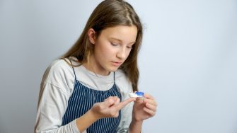 Portrait of positive cheerful little girl care eye hold transparent contact lens isolated over gray color background.