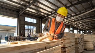 Worker wearing mask inspects wood in warehouse.