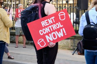 A person holds a placard in a protest gathering with the words "rent control now"
