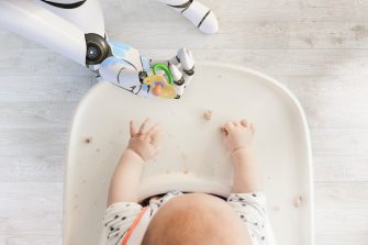 Robot hand giving pacifier to baby boy sitting in high chair playing with bread crumps, top view
