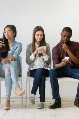 Diverse millennial  people waiting in queue sitting on chairs using devices