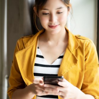 Asian woman standing side window with beautiful smile and using application chatting on cell smartphone while relaxing indoors