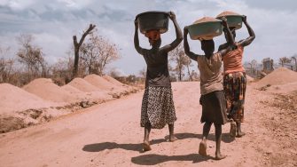 Two women and a boy holding a bag of soil on their head and walking barefoot