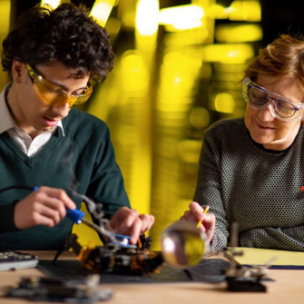 Founding Dean, Professor Judy Raper (R), wears lab goggles and holds a yellow pencil and works alongside a male undergraduate student (L) who wears yellow lab goggles while soldering electronics of a small robotic device on a work bench.