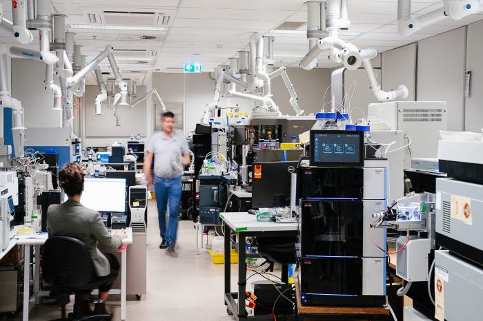 mass spectrometry lab with lots of analytical instruments that look like black and grey boxes and venting arms coming down from the ceiling. One staff member sits at a desk while the other is walking toward the camera.