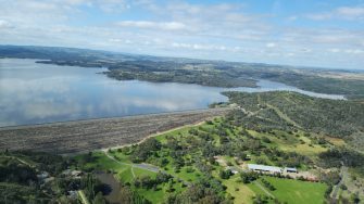 Aerial photo of an inland dam and the wall of the dam. The water is very high in the dam. The landscape around is green and vegetated with some cleared spaces, roads and buildings in the foreground.  The water surface is reflecting the blue cloudy sky.