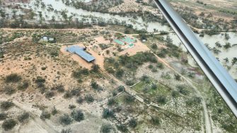 Aerial photo of shearing shed and quarters surrounded by flood waters. The river is running in the background. There ivegetation on the floodplain is green. There are a few dirt roads coming to the buildings.