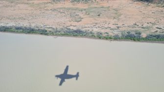 aerial photo of rive with brown water. Orange desert at river bank. Landscape covered by low green desert vegetation. Plane shadow on water in foreground