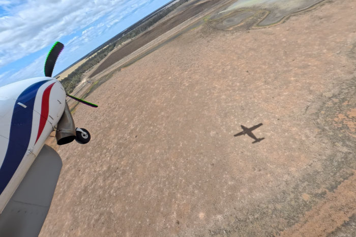 View of front section of light plane over dry red/brown earth. Shadow of plane visible on ground