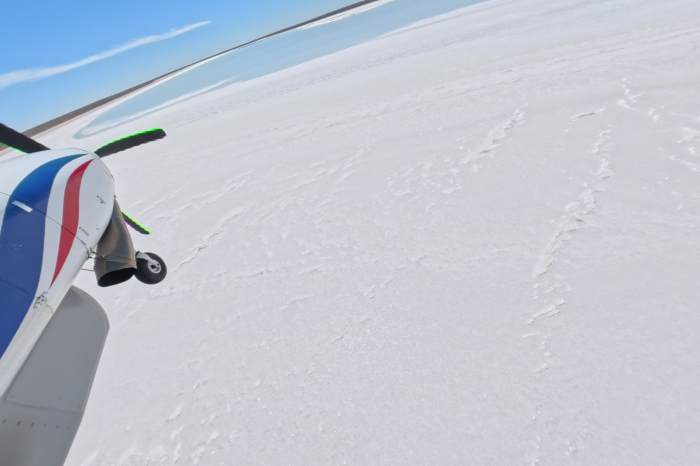 View of front section of light plane over white earth with blue water, shore line and sky in distance 