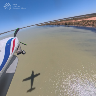 View of front section of light plane over water with red earth and some green vegetation at right