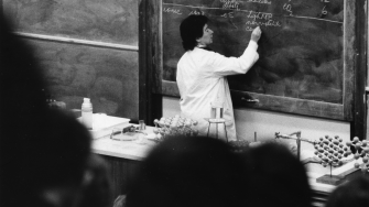 Dr June Griffiths standing at the front of a lecture hall writing on a blackboard