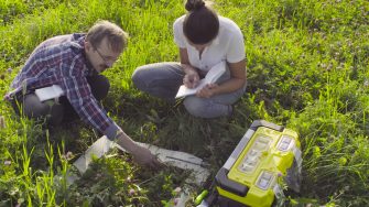 Crane shot, top view. Two ecologist examining plants on the meadow. Woman writing something in the diary. Field work