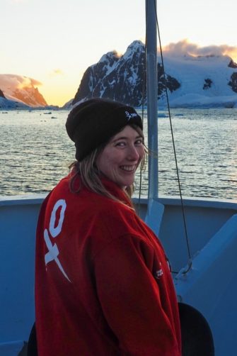 An image of a woman smiling at the camera from the bow of a boat with antarctic ice in the background.