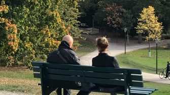 Two people sitting on a green bench with trees in the background