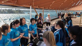 Group of students wearing blue tshirts near the water, with a table with laptops