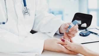 Stock image of person in white labcoat with stethoscope performing finger prick blood test on female patient