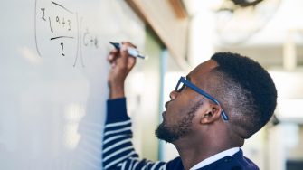 Man with glasses in striped jumper writing mathematical equation on whiteboard