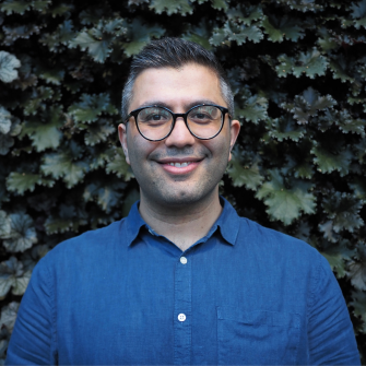 Portrait image of Dr Omid Ghasemi wearing a blue shirt and black framed optical glasses stnading infront of a green wall of dark green leaves