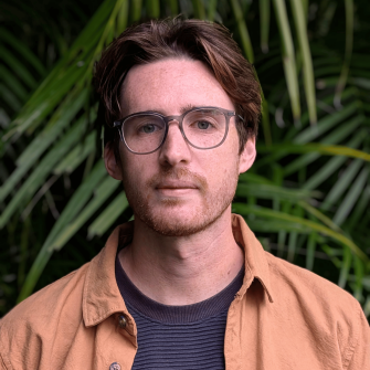 Portait image of Zac Coates wearing grey framed optical glasses standing in front of a green wall
