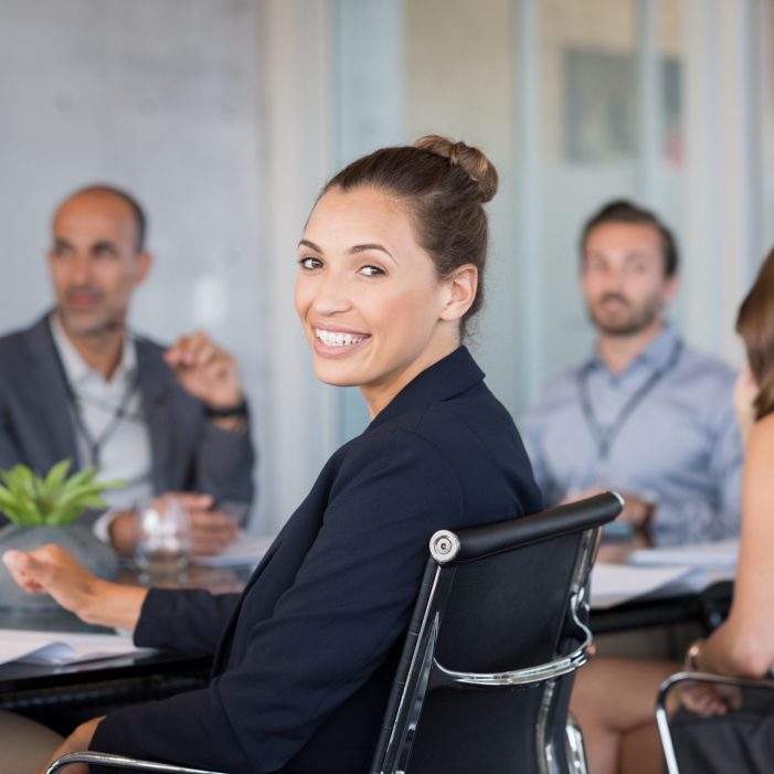 Young beautiful businesswoman with colleagues sitting in a modern board room. Proud smiling business woman sitting during a meeting and looking at camera. Portrait of a happy businesswoman with executives working.