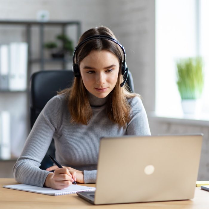 Beautiful young girl studying online using laptop web camera, writing lecture in notebook.Online education, remote working, home education.Online meeting,video call, video conference, courses online.