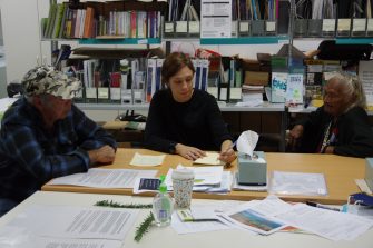 Three people, two older and young younger, are sitting at a table, looking at a notebook that one is writing in