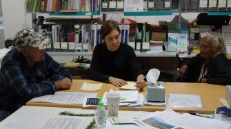 Three people, two older and young younger, are sitting at a table, looking at a notebook that one is writing in