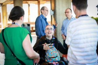 A small group of people gather indoors around a wheelchair user, talking together near concrete pillars with soft daylight filtering through windows.