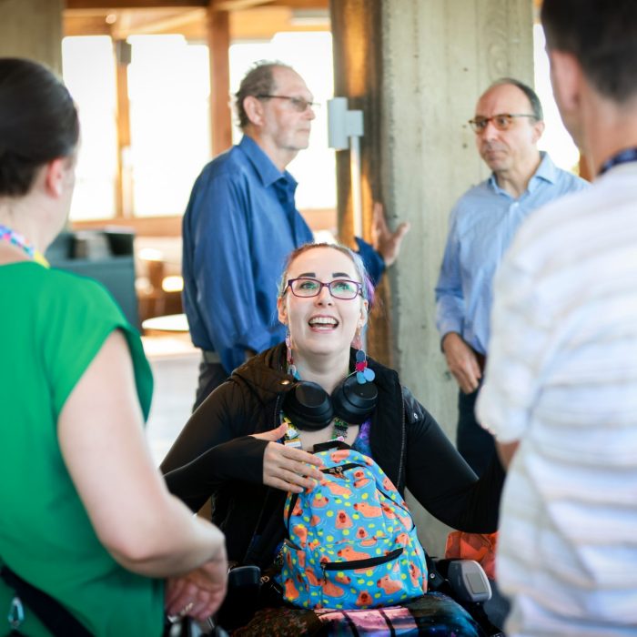 A small group of people gather indoors around a wheelchair user, talking together near concrete pillars with soft daylight filtering through windows.