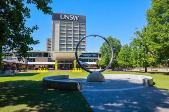 A circular metal sculpture stands on a paved lawn area with trees and the UNSW Main Library building visible in the background.
